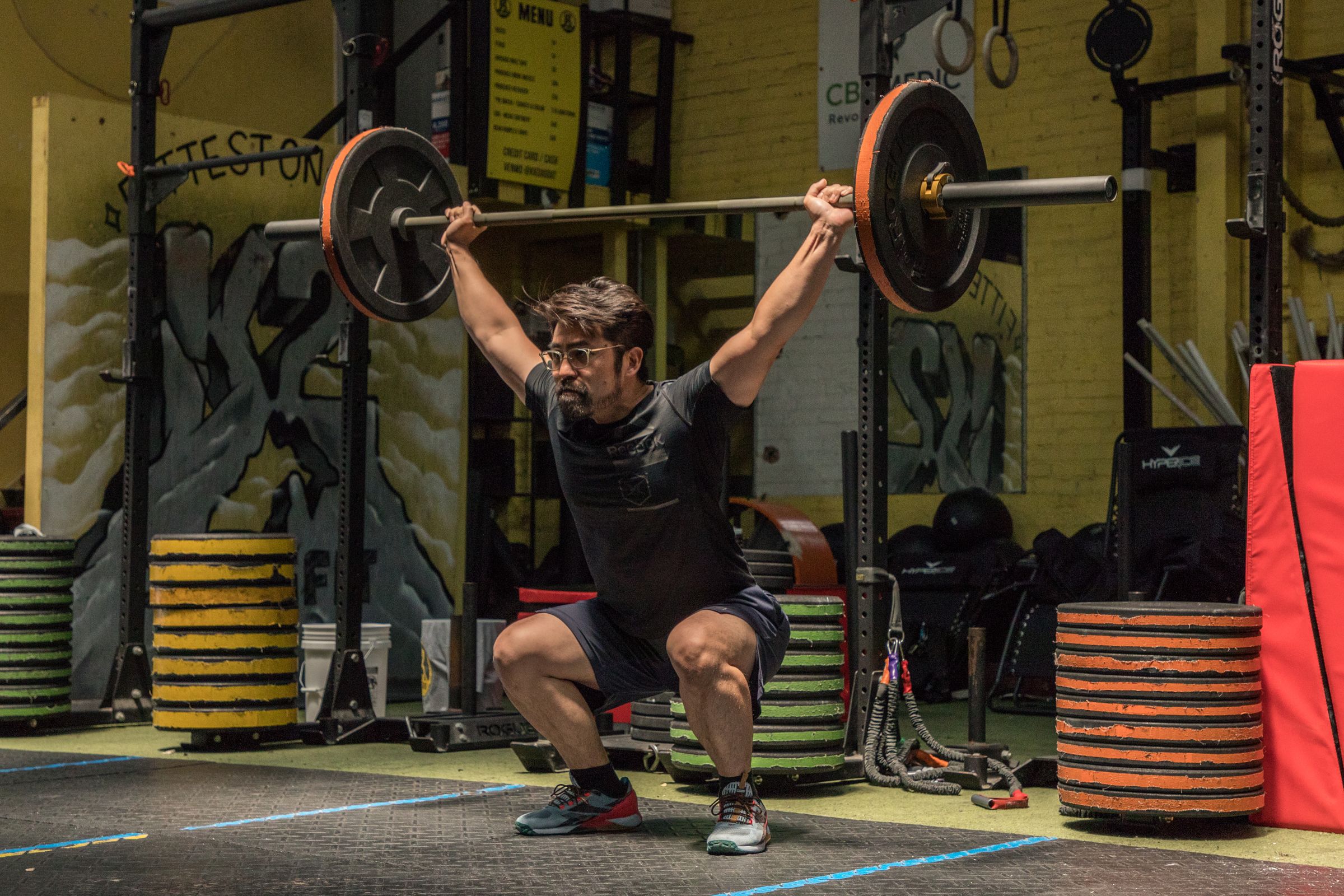 Coach Kevin Kim mid overhead squat in front of the yellow K2 mural at K2 HYROX Koreatown